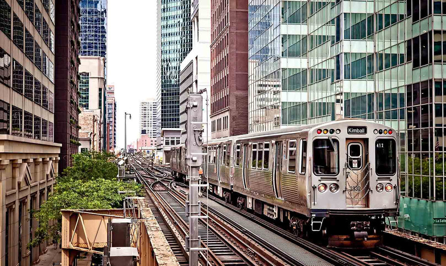 A modern conference room with a long wooden table, black chairs, and glass walls. The back wall displays the "Train Moving On The Tracks Wall Mural," showing elevated train tracks, tall buildings, and a train passing through the city.