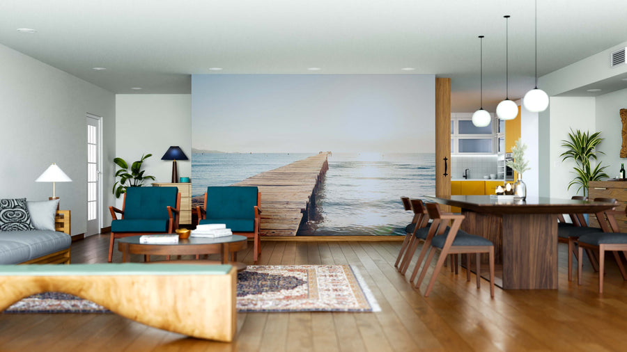 A modern, open living and dining area with wooden floors, teal chairs, and a large table is brightened by plants and the Tranquil Glow Wall Mural—a serene view of a wooden pier under a blue sky—while natural light fills the space.