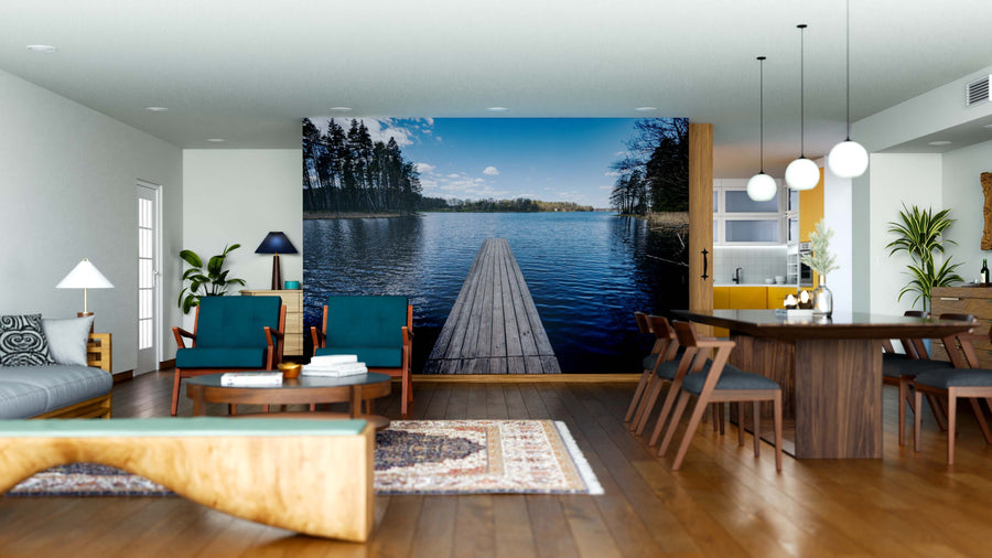 A modern open-plan living and dining area with wooden floors, teal and wood furniture, and a Tranquil Waters Wall Mural featuring a dock over a calm lake under blue skies, creating a serene and spacious atmosphere.