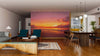 A modern living-dining room with wooden floors, teal armchairs, a wooden table, and spherical pendant lights features the Tropical Beach Bali Wall Mural, showcasing a vivid orange and purple beach sunset as a striking focal point.