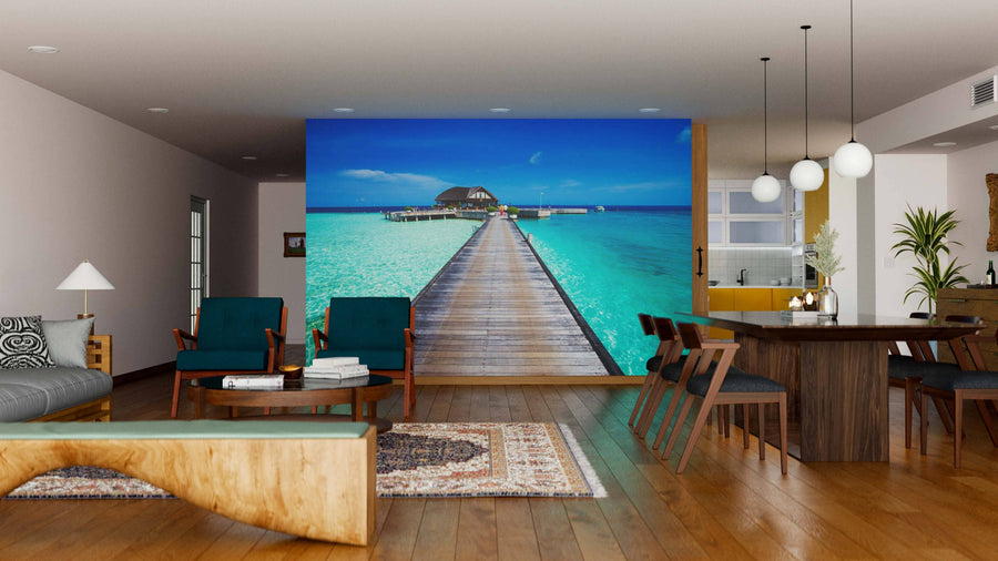 A modern living-dining room with wood floors and mid-century furniture features the Tropical Beach in Maldives Wall Mural, depicting a pier over turquoise water. Spherical pendant lights above a dark dining table enhance the inviting, tropical vibe.