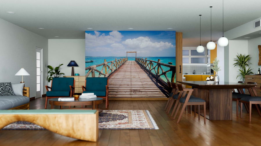 A modern living and dining area with wood furniture, blue chairs, rugs, and pendant lights feels open and tropical with the Tropical Pier Escape Wall Mural depicting a wooden pier over turquoise water under a sunny sky.