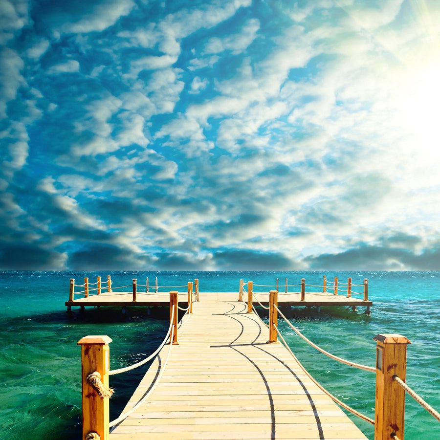 A modern open-plan living and dining room with wooden floors, teal and wood furniture, and a kitchen features the Tropical Wooden Pier Wall Mural—a vibrant peel-and-stick mural showing a pier leading to the ocean under a blue sky.
