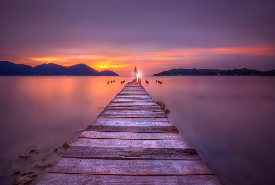 A modern living and dining space with teal chairs, wood furniture, and pendant lights features the Twilight Dock Wall Mural, showing a wooden pier stretching into a calm lake at sunset beneath a colorful mountain sky.