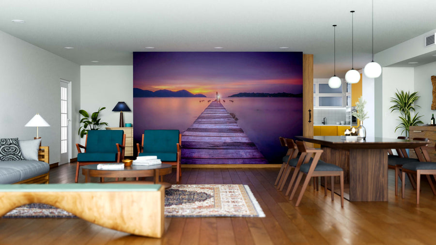 A modern living and dining space with teal chairs, wood furniture, and pendant lights features the Twilight Dock Wall Mural, showing a wooden pier stretching into a calm lake at sunset beneath a colorful mountain sky.