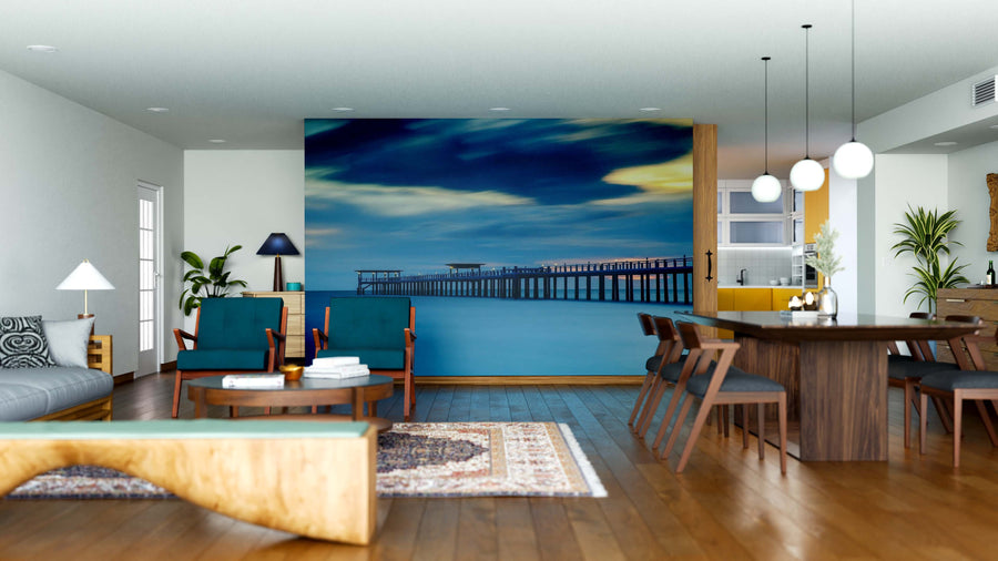 A modern open-plan living and dining room with wood floors, teal and wood furniture, pendant lamps, plants, and a feature wall displaying the Twilight Ocean Pier Wall Mural of a pier stretching into the ocean under a dramatic sky.