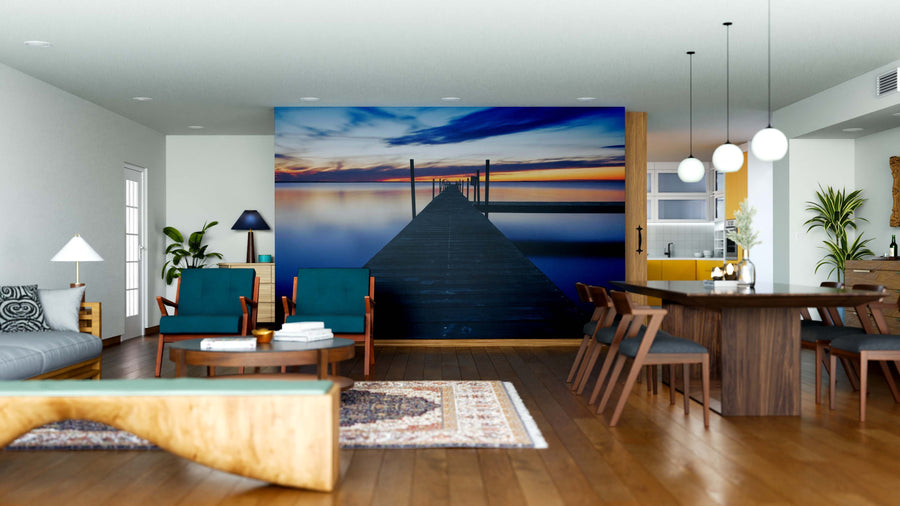 A modern open-concept living and dining room features wood floors, teal chairs, the Twilight Pathway Wall Mural of a dock at sunset, a long wooden dining table with chairs, pendant lights, and contemporary-rustic decor.