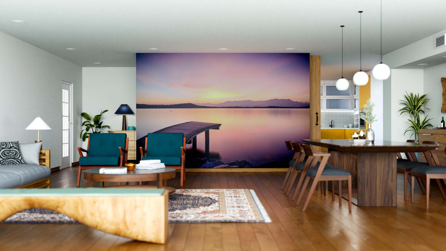 A modern open-plan living and dining room features wood furniture, rugs, green and blue accents, and plants. The back wall showcases the Twilight Rest Wall Mural depicting a serene lake at sunset, creating a peaceful focal point.