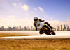 A motorcyclist in a white helmet and suit leans into a turn on a paved road. The predominantly white and yellow bike mirrors the sleek design of the Urban Speed Chase Wall Mural. A city skyline is visible under the clear blue sky, with dry grass lining the roadside in front.