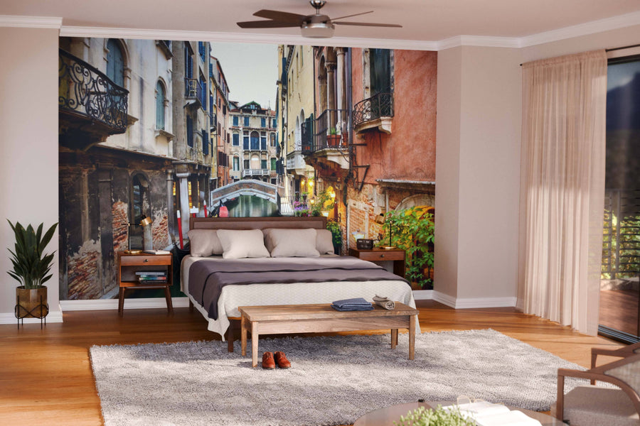 A cozy bedroom with beige walls features the Venice Canals Wall Mural behind the bed, complemented by wood furniture, potted plants, a fuzzy gray rug, and sheer curtains that let in natural light from wide windows.