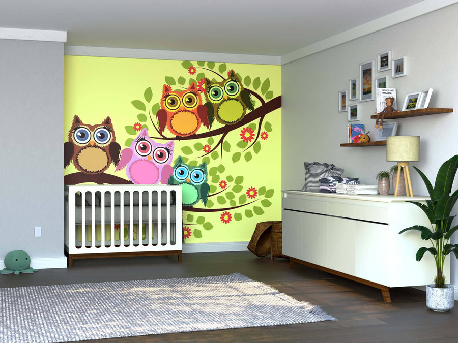 A nursery room features the Vibrant Owl Family Wall Mural—five colorful cartoon owls on a tree branch above a white crib. A dresser, framed photos, plant, and hardwood floors add to the room’s bright and playful atmosphere.