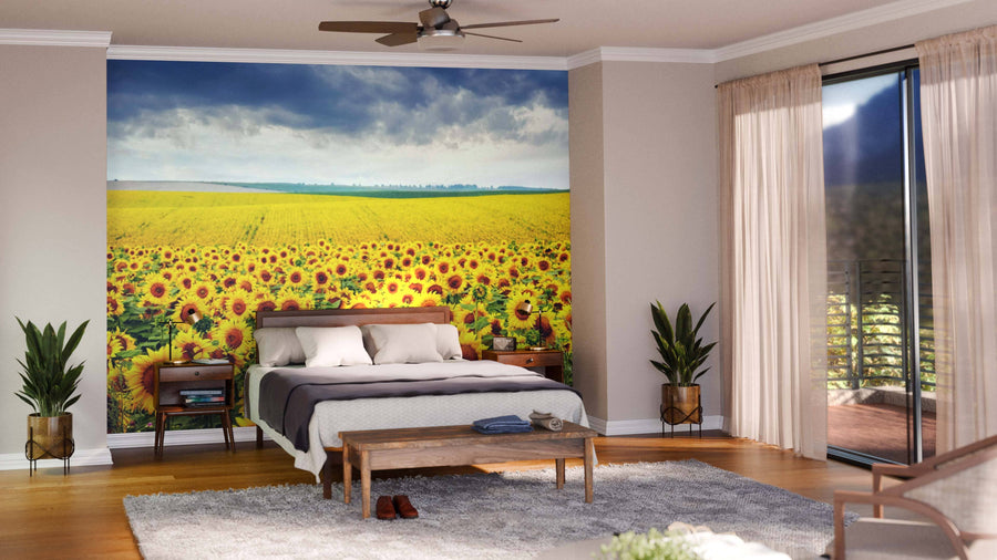 A modern bedroom showcases the Vibrant Sunflower Field Wall Mural behind a bed with gray and white bedding, flanked by bedside tables holding lamps and potted plants. Large windows, sheer curtains, and wooden accents enhance the space.