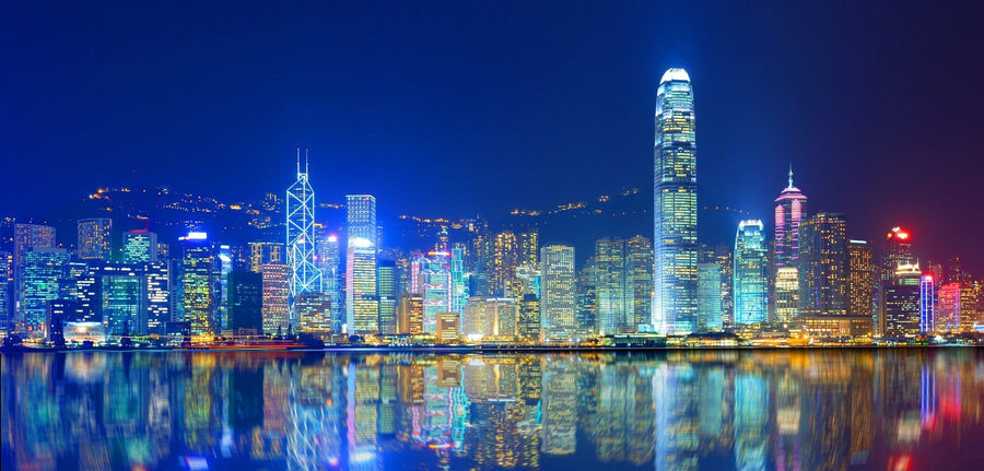 A modern living and dining space with wooden floors and furniture features the Victoria Harbor Wall Mural, displaying vibrant nighttime skyscrapers reflected on water. Warm hanging lights and rugs complete this inviting, stylish room.