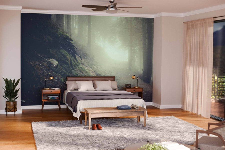A modern bedroom with a Vintage Mountain Foggy Forest Wall Mural behind a wooden bed, light bedding, two mid-century nightstands and lamps, a foot bench, potted plant, large rug, wood floors, beige walls, and sheer curtains letting in sunlight.