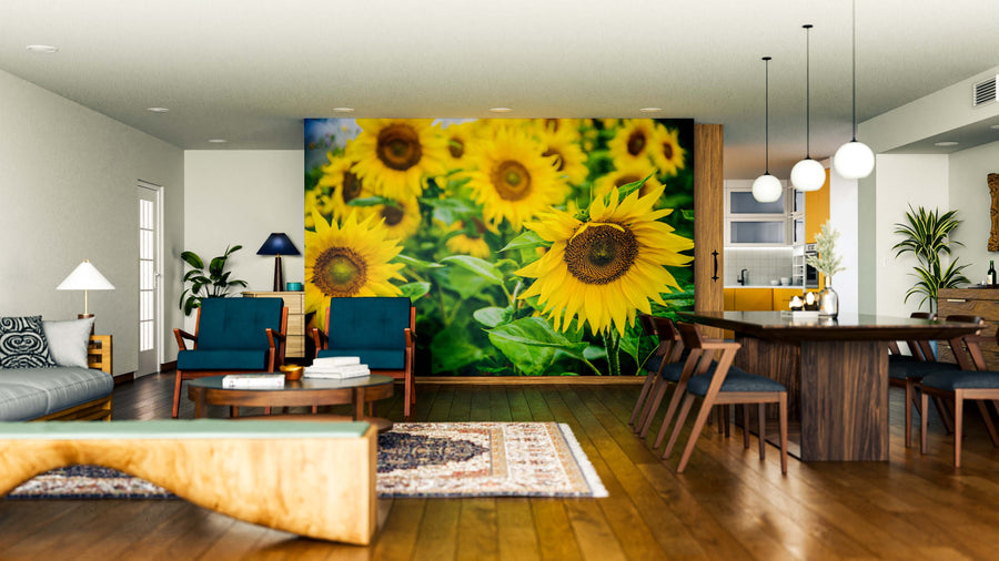 A bright living-dining area features the Warm Summer Field Wall Mural of sunflowers, wood floors, modern dark blue chairs, a wooden table, pendant lights, green plants, a rug, and ample natural light for a fresh, cozy feel.