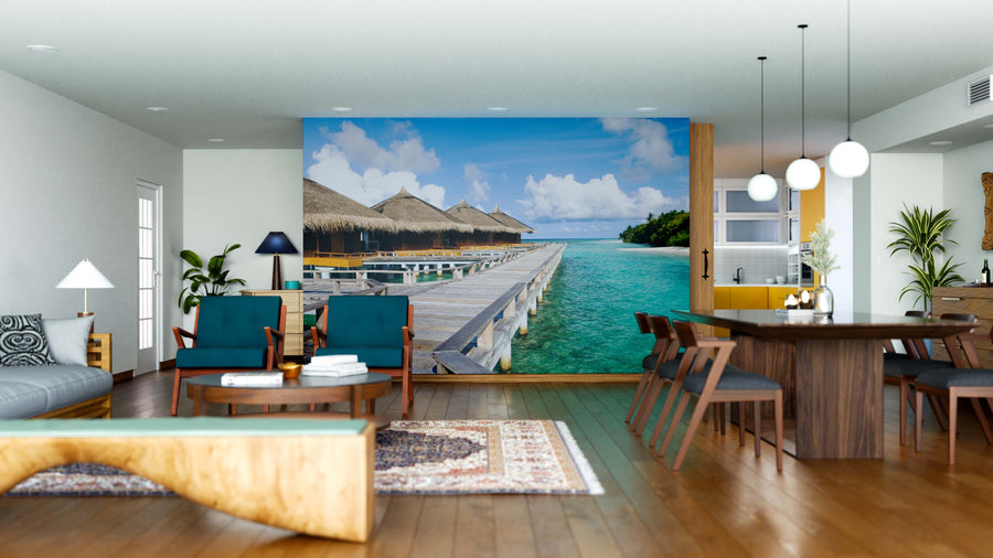 A modern living-dining area features wood furniture, blue armchairs, pendant lights, and the Water Villas Wall Mural depicting an overwater bungalow walkway above turquoise water for a tropical vibe. Rugs and decor add warmth to the space.
