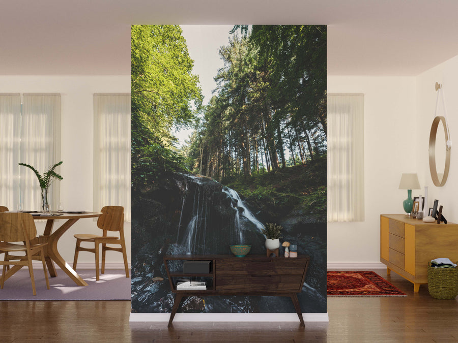A living room with a dining area features the Water falling on Wet Stones Wall Mural. Wooden furniture, plants, a round mirror above a dresser, and soft natural light from tall windows with sheer curtains complete the scene.