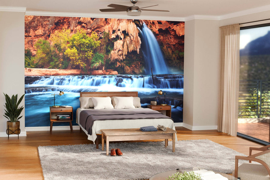 A modern bedroom with wooden furniture and large windows features the Waterfall Wall Mural, showcasing a vibrant waterfall and lush greenery to create a serene, nature-inspired space filled with natural light and effortless style.