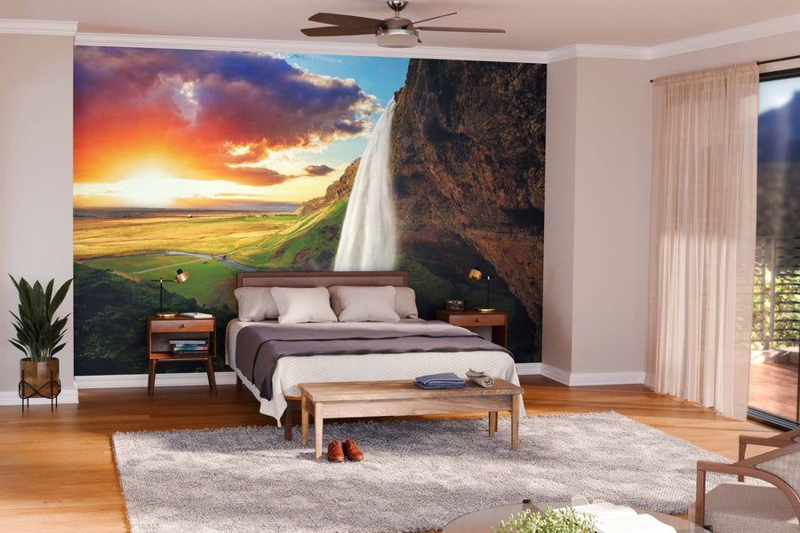A modern bedroom with the "Waterfall in Iceland Wall Mural" showcases a vivid landscape of cascading water, lush fields, and a sunset. A wooden bed, side tables, greenery, and sheer curtains complete the bright, inviting space.