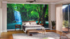 A modern bedroom with a bed, bench, rug, and potted plants features the "Waterfall in Thailand National Park Wall Mural," creating an illusion of opening into nature. Sunlight streams through sheer curtains covering glass doors.