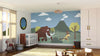 A nursery featuring the Wilderness Run Wall Mural, depicting a cartoon mammoth chasing a person with a spear among hills, trees, and a volcano. The room includes a crib, toys, rocking horse, round mirror, and window with beige curtains.
