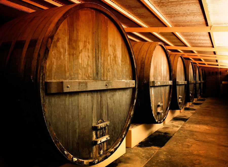 A stylish cafe with wooden tables and black chairs features the Winery Barrels Wall Mural. Pendant lights hang above, potted plants decorate tables, wine bottles line shelves, and large windows welcome natural light.