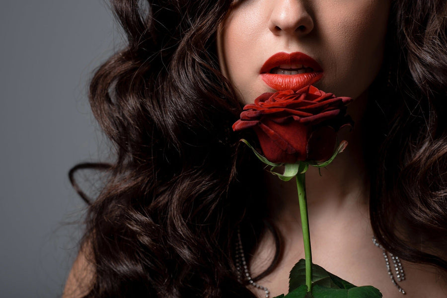 A woman with long, wavy dark hair and red lipstick holds a deep red rose near her lips in the Woman Holding Rose Wall Mural—perfect inspiration for custom printed murals or removable wallpaper. She wears a pearl necklace against a neutral gray background.