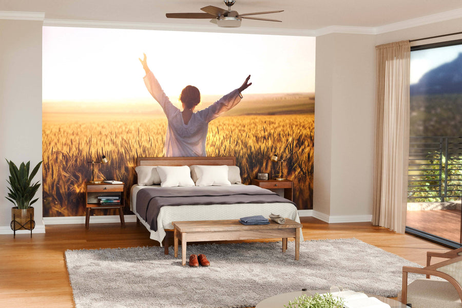 A bright bedroom features the Woman with Arms Outstretched Wall Mural behind the bed, showing a figure in a wheat field. The space includes wooden floors, double bed, nightstands, bench, plant, and large windows with sheer curtains.