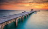 A tranquil Wood Bridge Wall Mural from Deposit Photo features a pier stretching over calm turquoise waters toward an open shelter, set against a sunset sky of orange, pink, and purple hues, casting a warm glow.