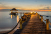 A tranquil scene depicted in the Wooden Bridge Landscape Wall Mural by features a wooden pier over calm water leading to a thatched-roof gazebo. The sunset sky's orange and blue hues, along with soft light on the pier, evoke serenity, while rocks enhance the natural beauty.