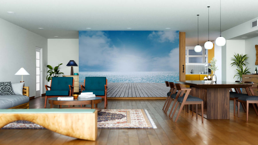 A modern living and dining room with wooden floors, blue and wood furniture, pendant lights, houseplants, rugs, and a “Wooden Dock With Ocean View Wall Mural” creates a warm, tranquil space featuring serene ocean scenery.