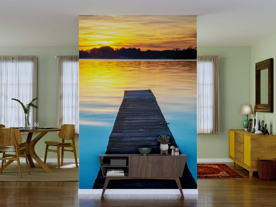 A living room with pale green walls and wood furniture showcases the Wooden Jetty Wall Mural, depicting a dock stretching into a serene lake at sunset, seamlessly blending nature’s beauty behind a mid-century console adorned with plants and decor.