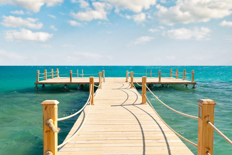 Spacious modern living and dining area with wood floors, teal chairs, and the Wooden Pier Wall Mural depicting a dock over turquoise ocean, creating an outdoor illusion. Eclectic decor, round lights, plants, and contemporary furniture accent the space.
