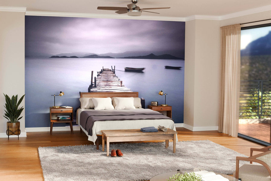 A serene bedroom features a wooden bed, beige and brown bedding, two nightstands with lamps, a bench, and the Wooden Pier with Boat Wall Mural behind the bed for a tranquil vibe. Large windows and neutral decor complete the calming space.