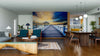 A modern living-dining room features wood furniture, teal chairs, and the Wooden Piers on Dusk Wall Mural as a serene focal point. Pendant lights, plants, and decor complete the inviting and airy space.