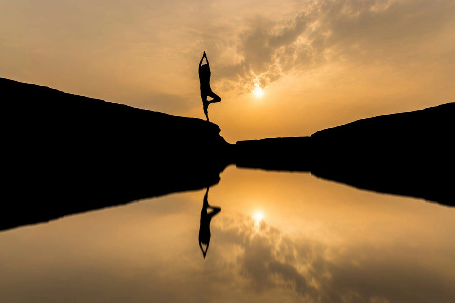 A modern living-dining room with wooden floors, stylish furniture, and a large Yoga Pose Wall Mural showing a person doing a handstand at sunset by a reflective lake. Round pendant lights hang above a wooden dining table for six.