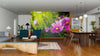 A modern living and dining room with wooden floors, dark blue chairs, a sofa, and houseplants features the Cosmos Flowers Wall Mural on the back wall—a vibrant display of pink cosmos blooms against a green, blurred background.