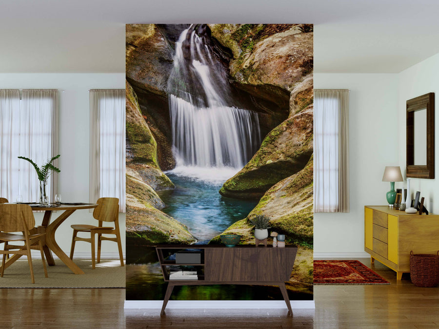 A living room features the Hocking Hills Splash Wall Mural with a waterfall and rocks as a partition. The space has a wooden dining table, yellow sideboard, modern decor, and sunlight streaming through sheer curtains for a cozy vibe.