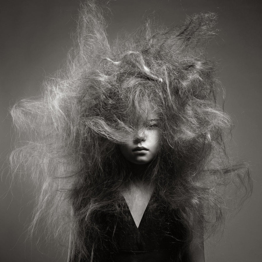 A person with a serious expression is partly obscured by voluminous, wild hair extending in every direction. Their deep V-neck contrasts with the bold hair texture. This black-and-white image vividly captures movement and resembles a custom printed mural. (Bedhead Wall Mural by BEAUTY).